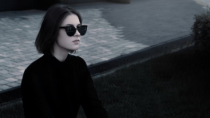 Young woman in black sunglasses and dark top sitting on a city street