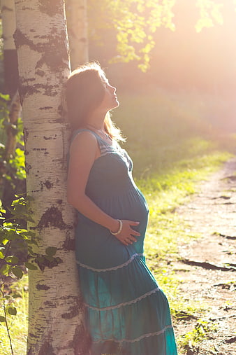 Pregnant woman in teal dress leaning against birch tree in sunlight