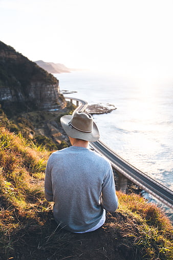 Man in Hat Sitting on Cliff Overlooking Coastal Bridge