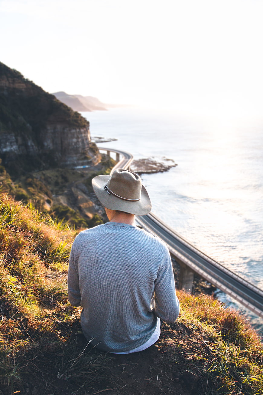 Man in Hat Sitting on Cliff Overlooking Coastal Bridge