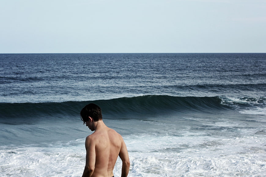 Shirtless Man Looking at Ocean Waves on Beach