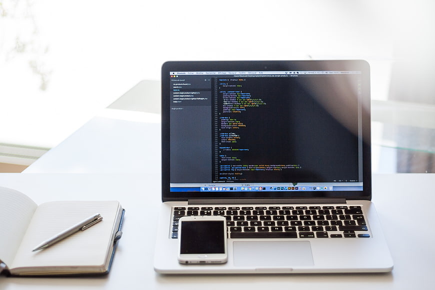 Laptop with code, smartphone, and notebook on white desk