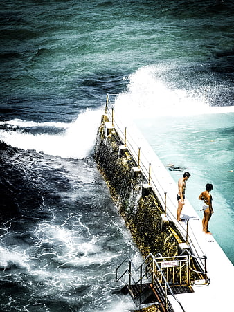 Couple standing on ocean pool edge watching crashing waves
