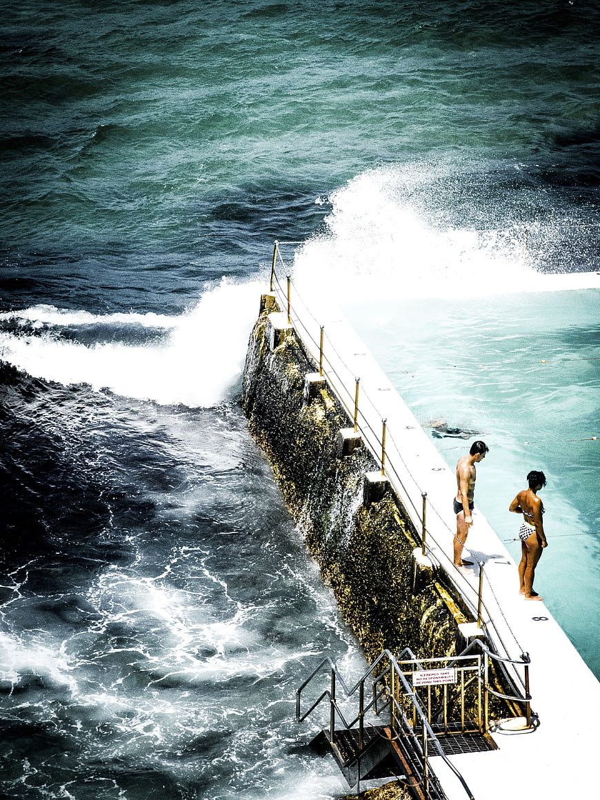 Couple standing on ocean pool edge watching crashing waves