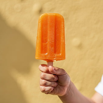 Hand Holding Orange Popsicle Against Yellow Wall