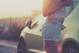 Young woman in denim shorts standing by car at sunset