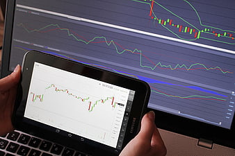 Hands holding tablet with stock market charts in front of laptop screen