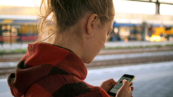 Woman in Red Plaid Hoodie Using Smartphone at Train Station