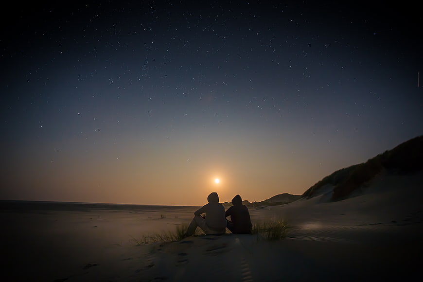 Couple watching moonrise over starry beach dunes