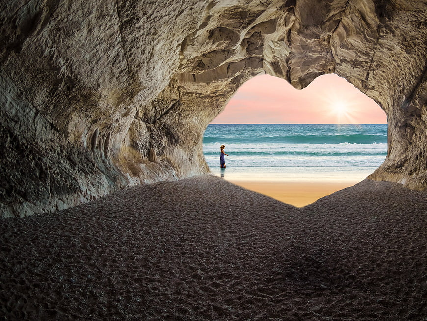 Heart-shaped cave opening to ocean sunset with woman