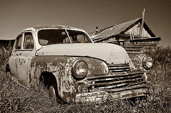 Abandoned vintage car in overgrown field with rustic cabin