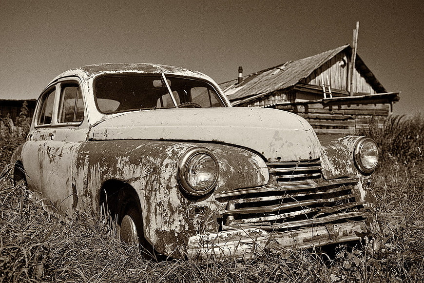 Abandoned vintage car in overgrown field with rustic cabin