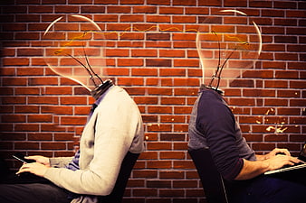 Two men with lightbulb heads working on laptops against a brick wall