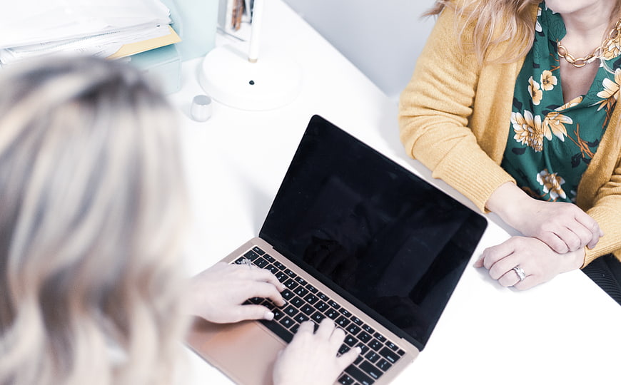 Two women collaborating at a white office desk with a laptop