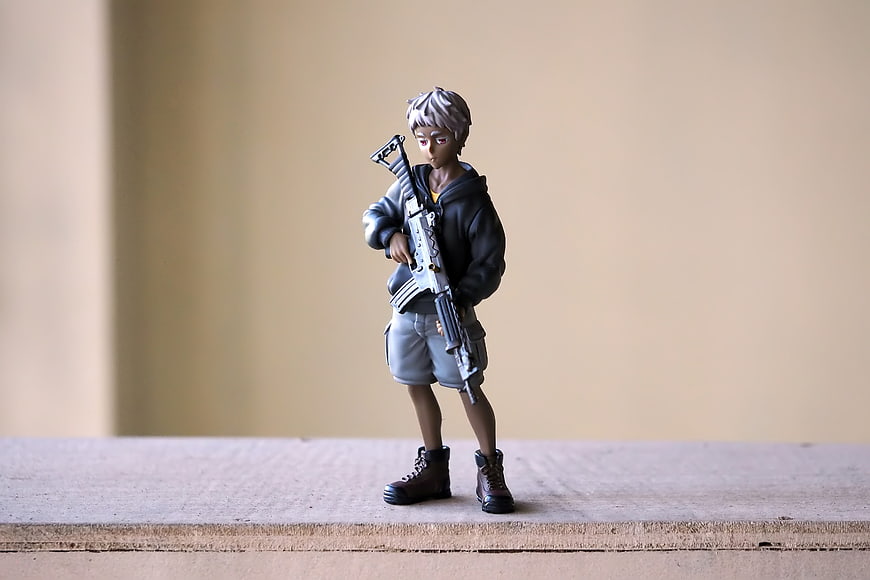 Anime Boy Figurine Holding Gun On Wooden Surface
