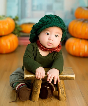 Cute baby in green knit hat sitting on wooden stool with pumpkins