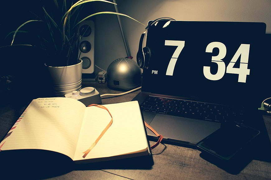 Laptop with Digital Clock and Open Notebook on Desk