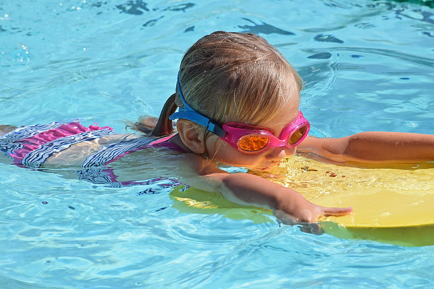 Young girl swimming with goggles and kickboard in pool