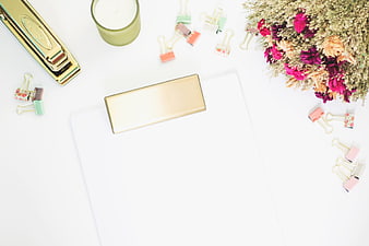 Flat lay of gold stapler, dried flowers, and pastel binder clips on white desk