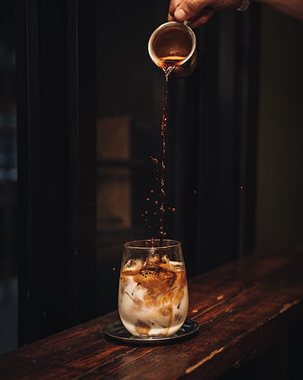 Barista pouring espresso into iced latte glass on dark wooden table