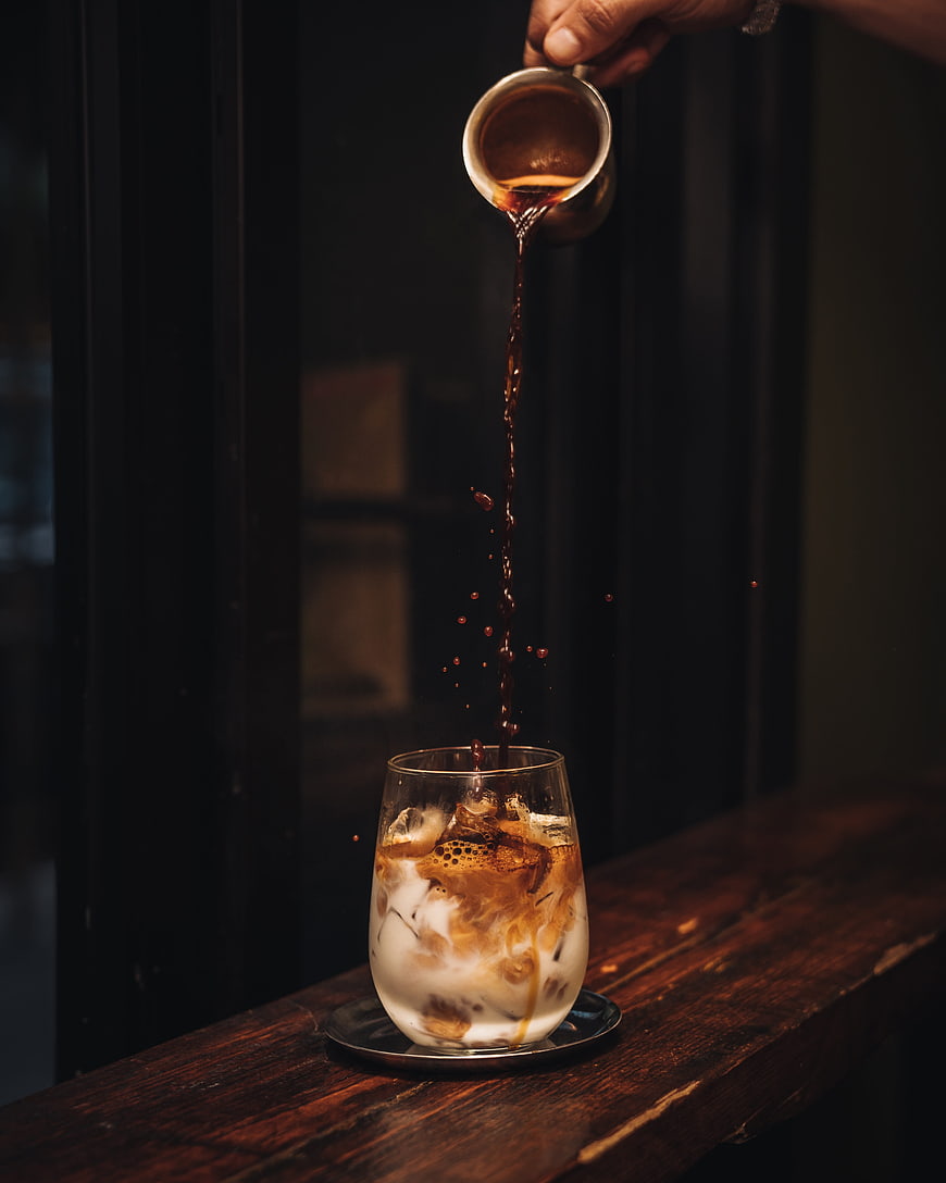 Barista pouring espresso into iced latte glass on dark wooden table
