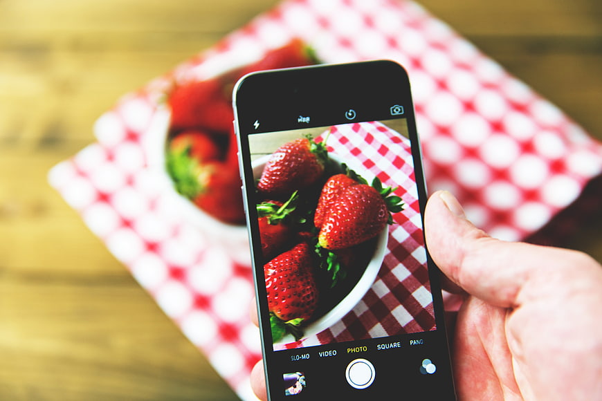 Hand holding smartphone photographing fresh strawberries on checkered cloth