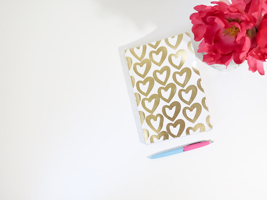 Flat lay of gold heart notebook with pink pen and red flowers