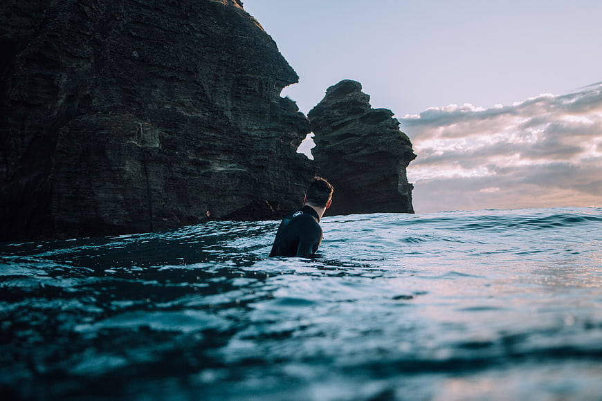 Surfer in Wetsuit Facing Rocky Cliffs at Sunset