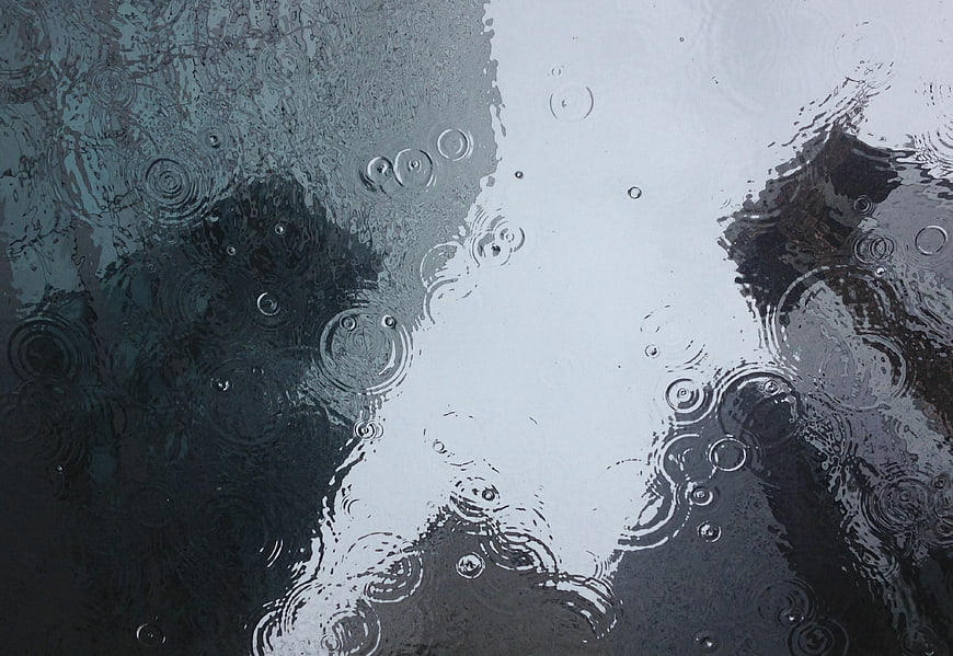 Raindrops creating ripples in a dark puddle with sky reflection