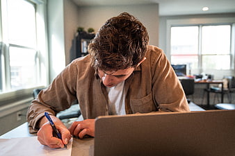 Young man writing notes at desk with laptop in office