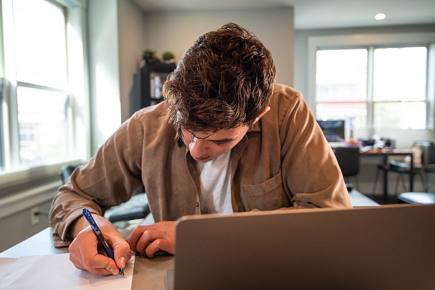 Young man writing notes at desk with laptop in office