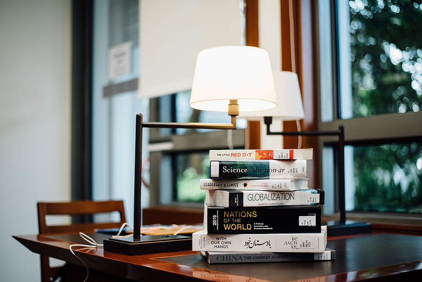 Stack of books and lamp on wooden desk by window