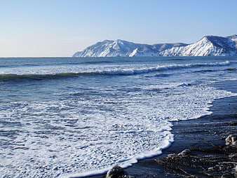Winter Ocean Waves Crashing on Dark Sandy Beach with Snowy Mountains