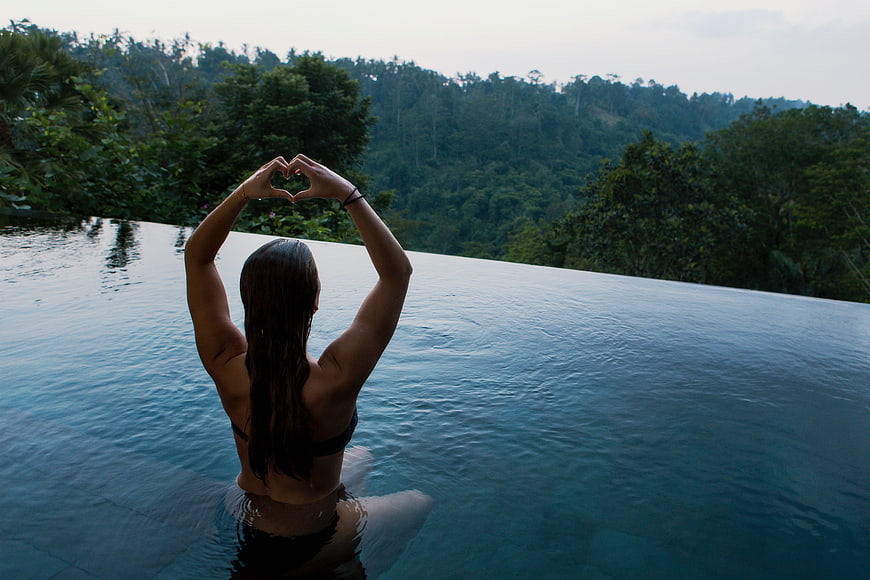 Woman making heart shape in infinity pool overlooking tropical forest