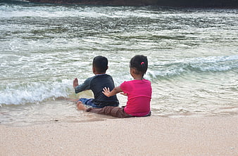 Two children sitting on sandy beach watching ocean waves