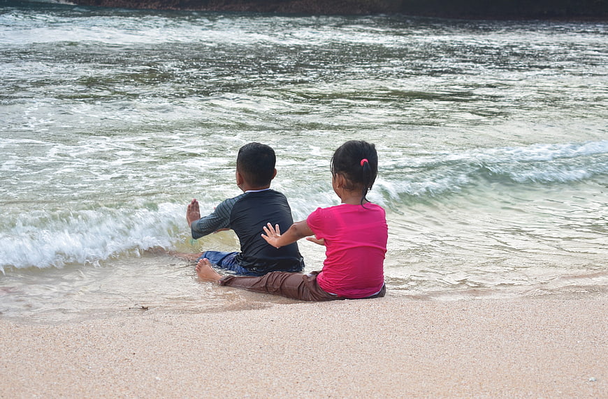Two children sitting on sandy beach watching ocean waves