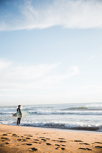 Surfer with board standing on sandy beach looking at ocean waves
