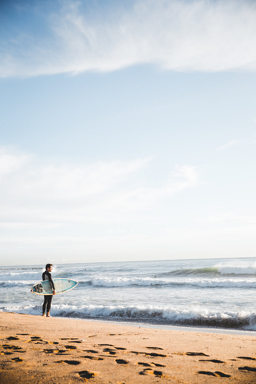 Surfer with board standing on sandy beach looking at ocean waves