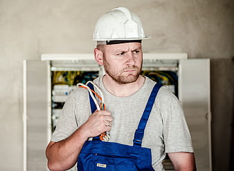 Focused electrician in blue overalls and white hard hat holding multimeter