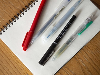 Assorted Pens on Spiral Notebook on Wooden Desk
