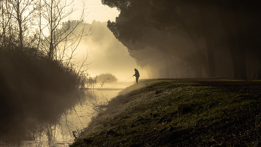 Silhouette of fisherman on misty riverbank at sunrise