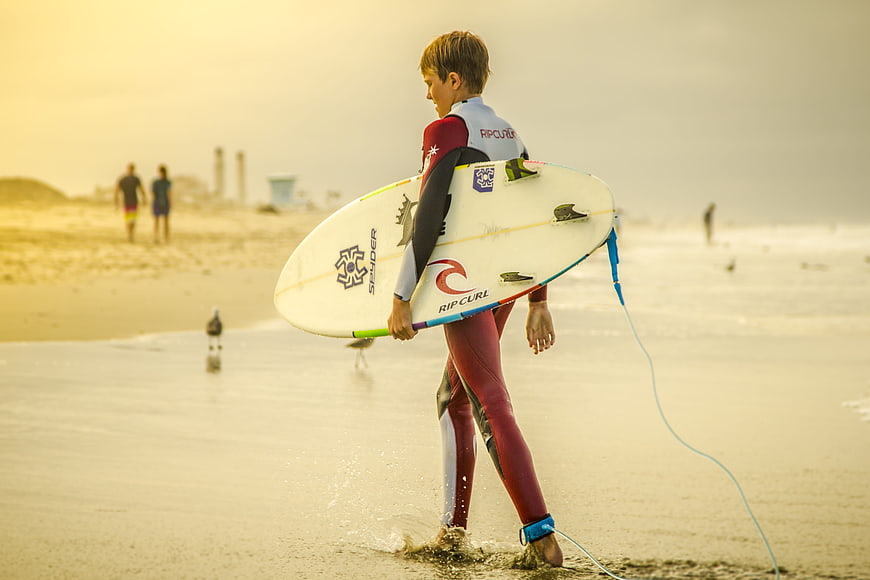 Young Surfer Walking on Beach with Surfboard at Sunset