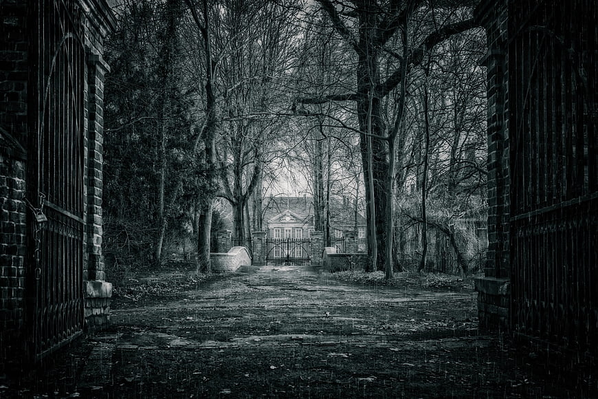 Dark moody black and white photo of an open iron gate leading to a house in a forest