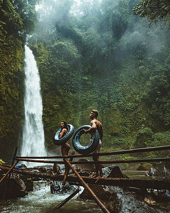 Couple with inner tubes on bamboo bridge near tropical waterfall