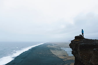 Man standing on cliff overlooking black sand beach and ocean