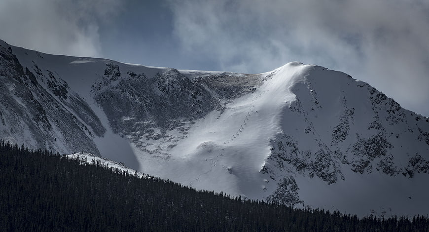 Snow-Covered Mountain Peak with Dark Forest and Cloudy Sky