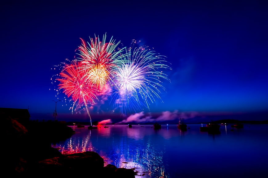 Colorful fireworks exploding over a calm harbor at night with boat silhouettes