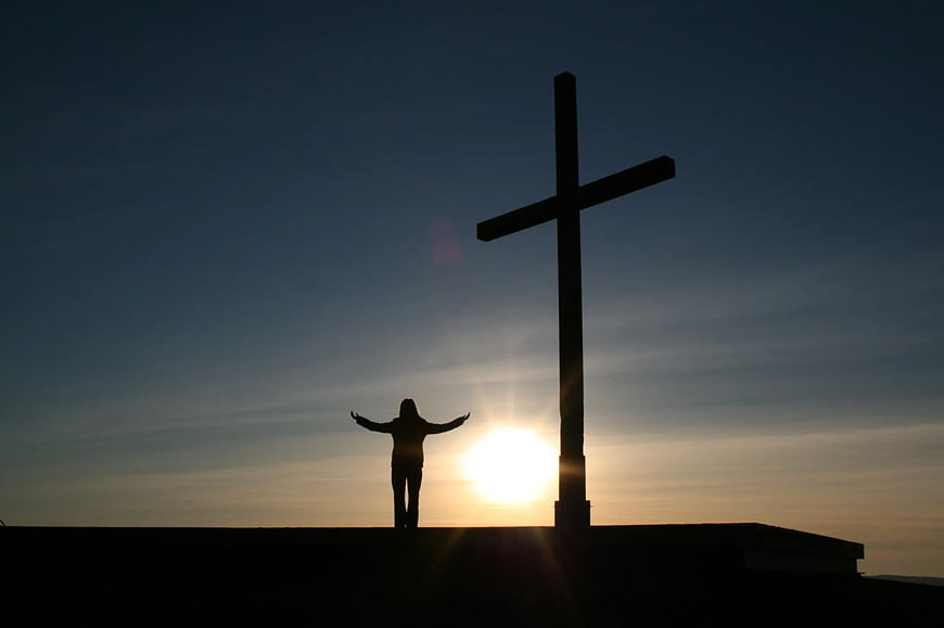 Silhouette of Person with Arms Raised Beside Cross at Sunset