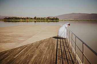 Bride in white gown leaning on pier railing by lake