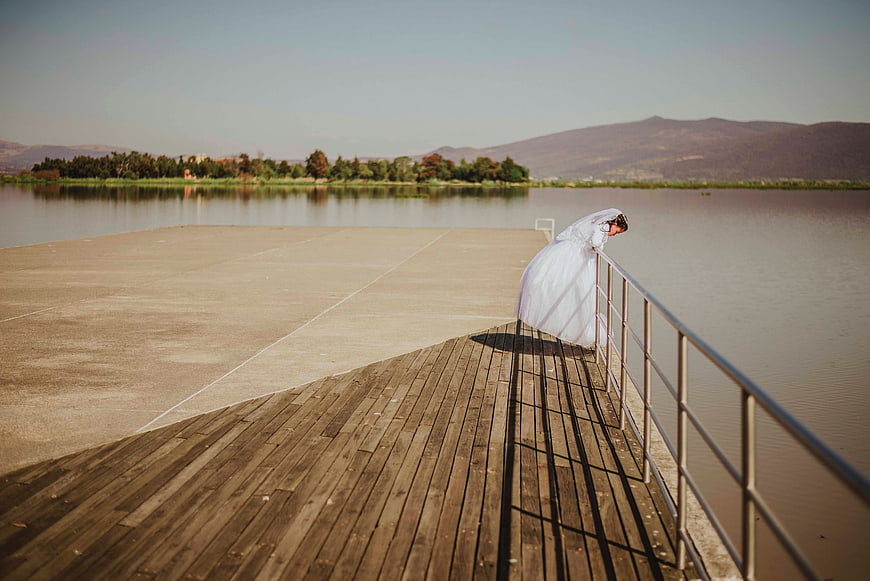 Bride in white gown leaning on pier railing by lake